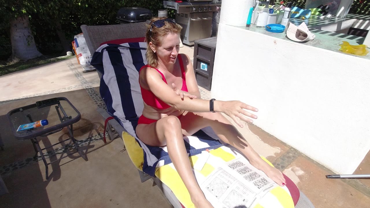 At 0:01 in Los Cabos, Mexico, the seated woman continues rubbing sunscreen onto her outstretched arm, with the white patio wall and small side table visible beside her.