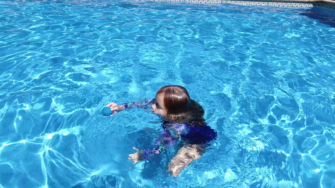 The child remains partly submerged in a Los Cabos pool, extending an arm toward the blue ball while sunlight creates sharp white patterns across the water.