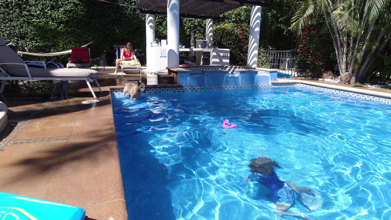 The Los Cabos pool scene holds wide, with a child swimming underwater near the foreground, a pink toy floating mid-pool, and adults seated by the shaded patio.