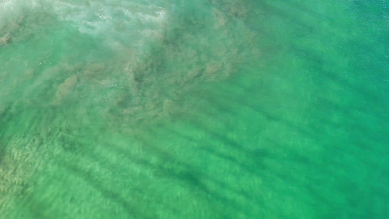 Overhead view of turquoise-green ocean water over pale underwater sand patterns in Los Cabos, Mexico, with no visible people.
