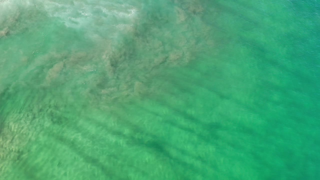 Turquoise ocean surface with darker underwater ridges running diagonally through the sandy bottom in Los Cabos, Mexico, with no visible people.