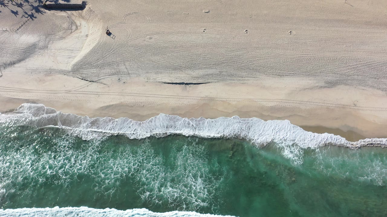 High aerial view in Los Cabos, Mexico, with green ocean water breaking into white foam along a wide pale beach and no visible people.