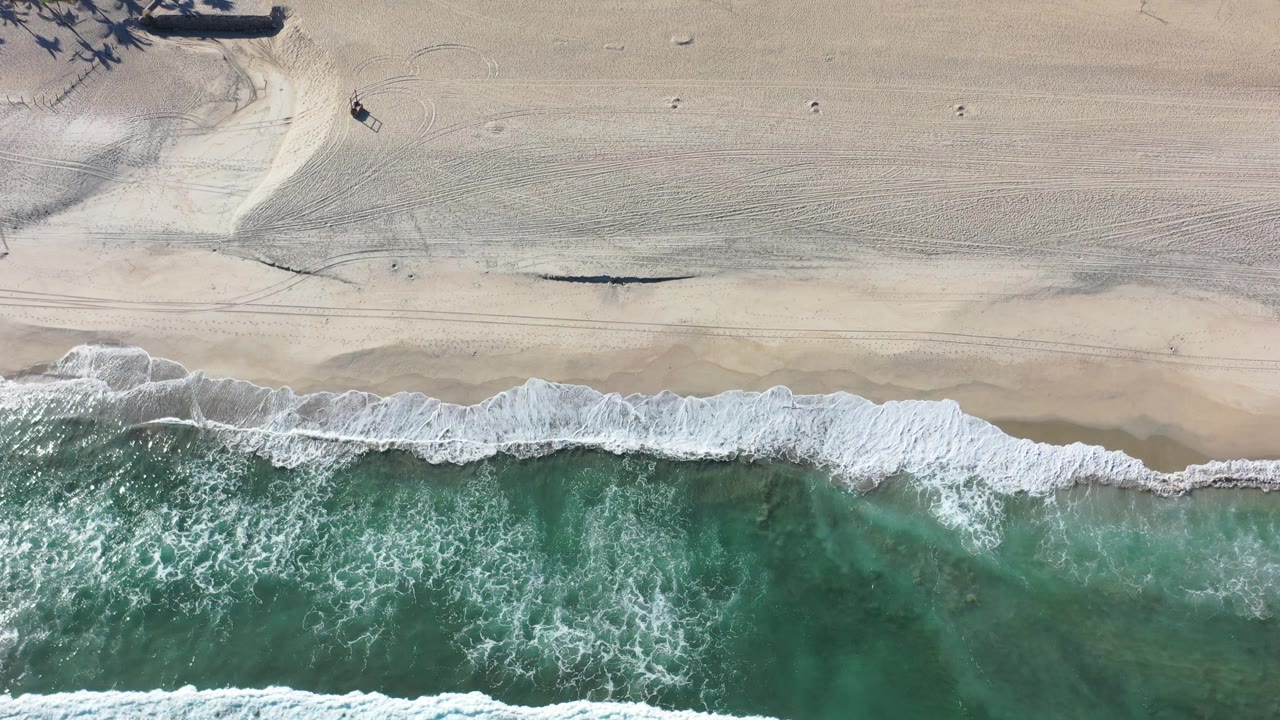 Drone-style overhead frame in Los Cabos, Mexico, showing foamy waves crossing the lower half of the image and broad raked sand above with no visible people.