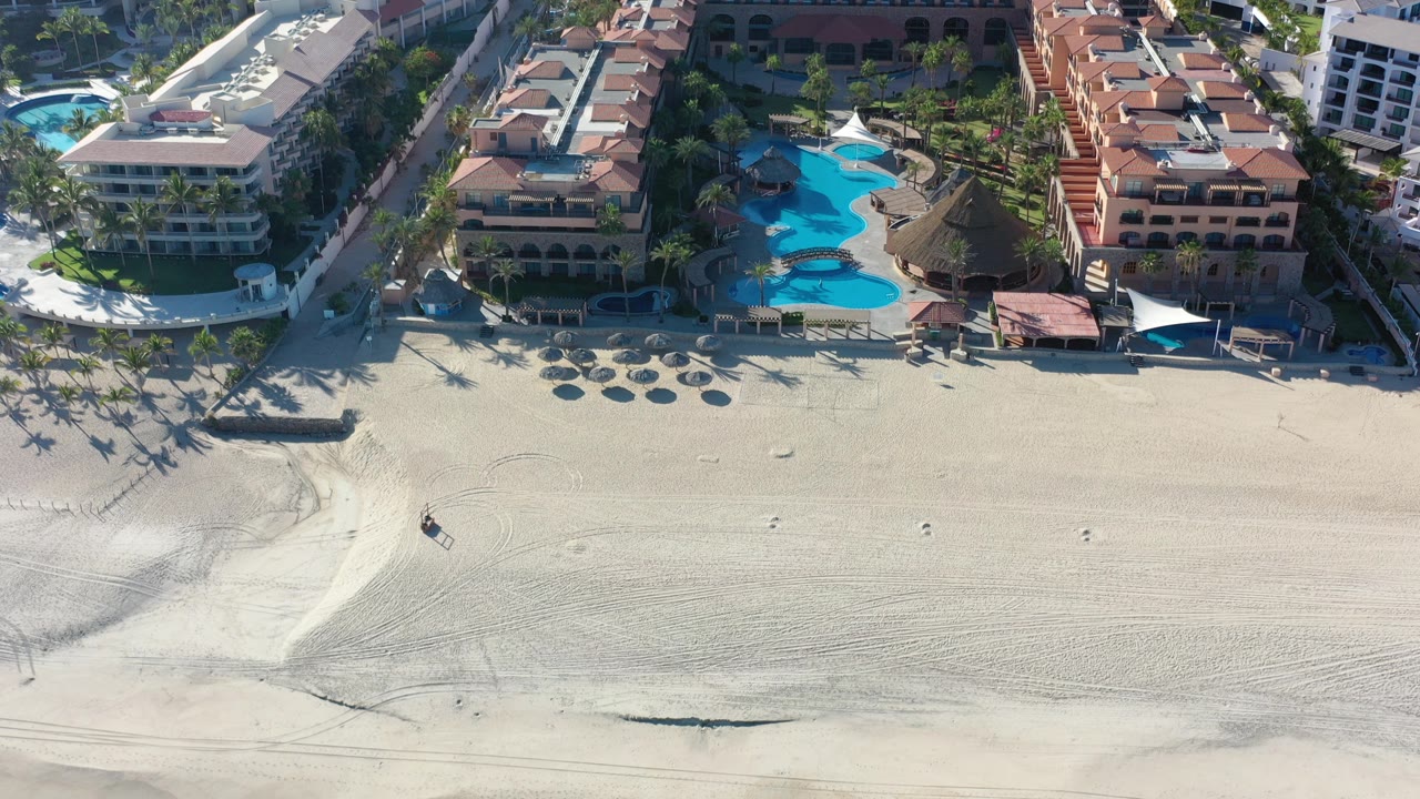 Aerial view of beachfront hotels, pools, palms, and pale sand.