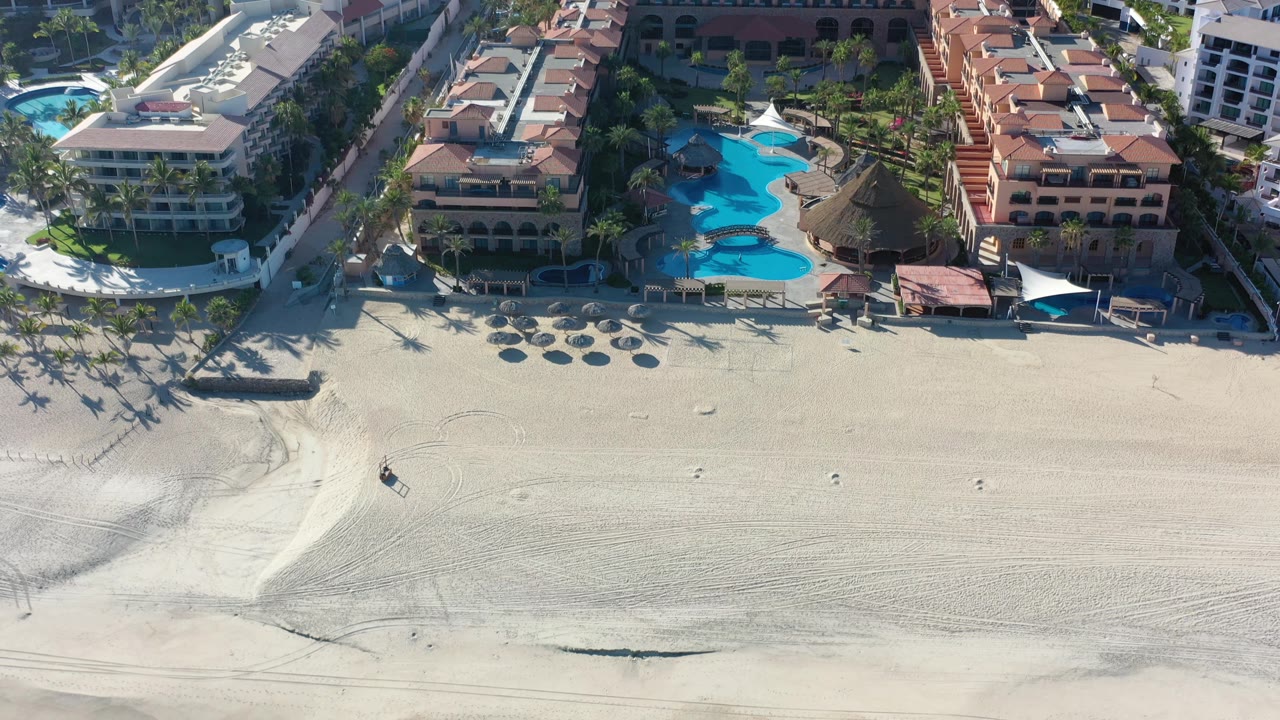 Aerial view in Los Cabos, Mexico, with the beach spanning the lower frame, palm shadows crossing the sand, and bright pools set between hotel buildings.