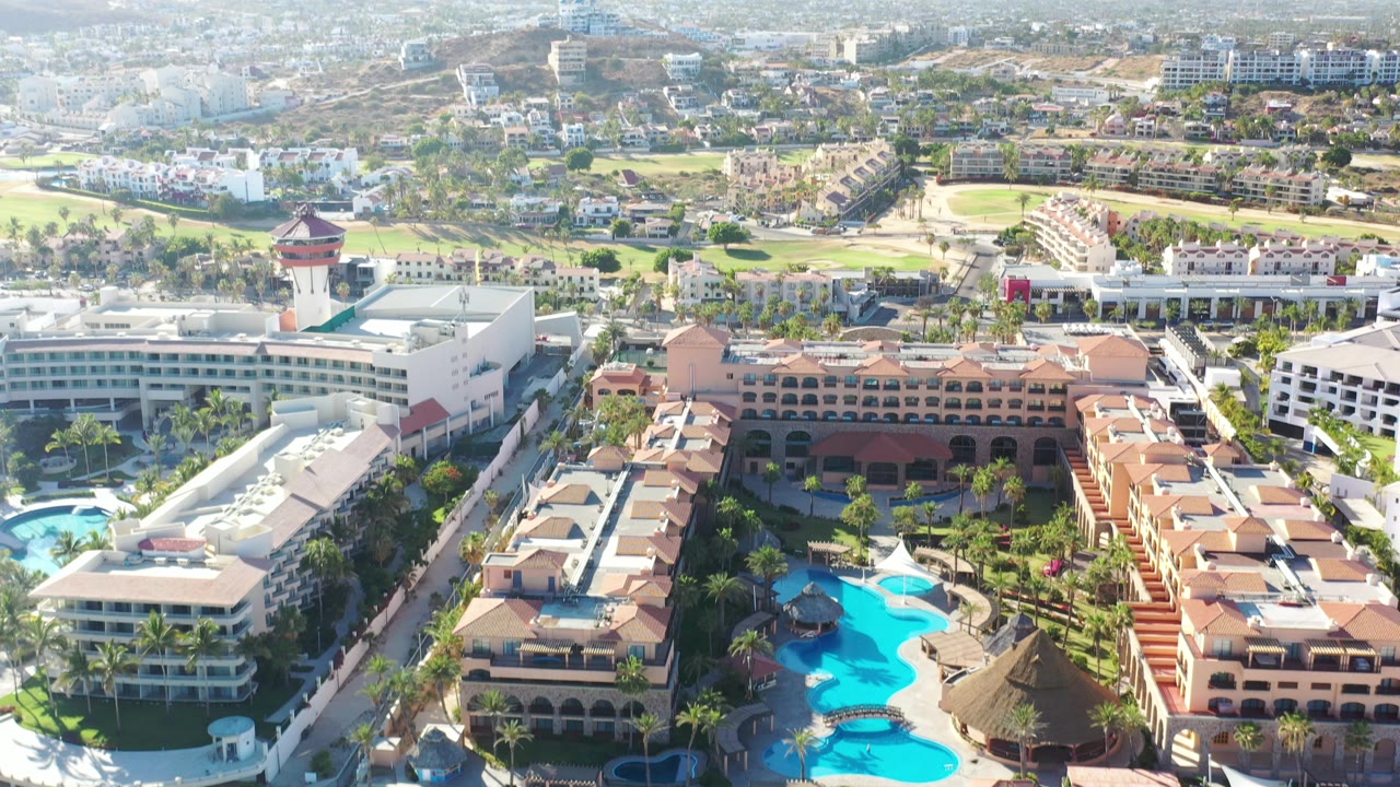 Opening aerial frame over Los Cabos, Mexico, with terracotta resort roofs, bright blue pools, palm landscaping, golf greens, surrounding development, hazy mountains, and no visible people.