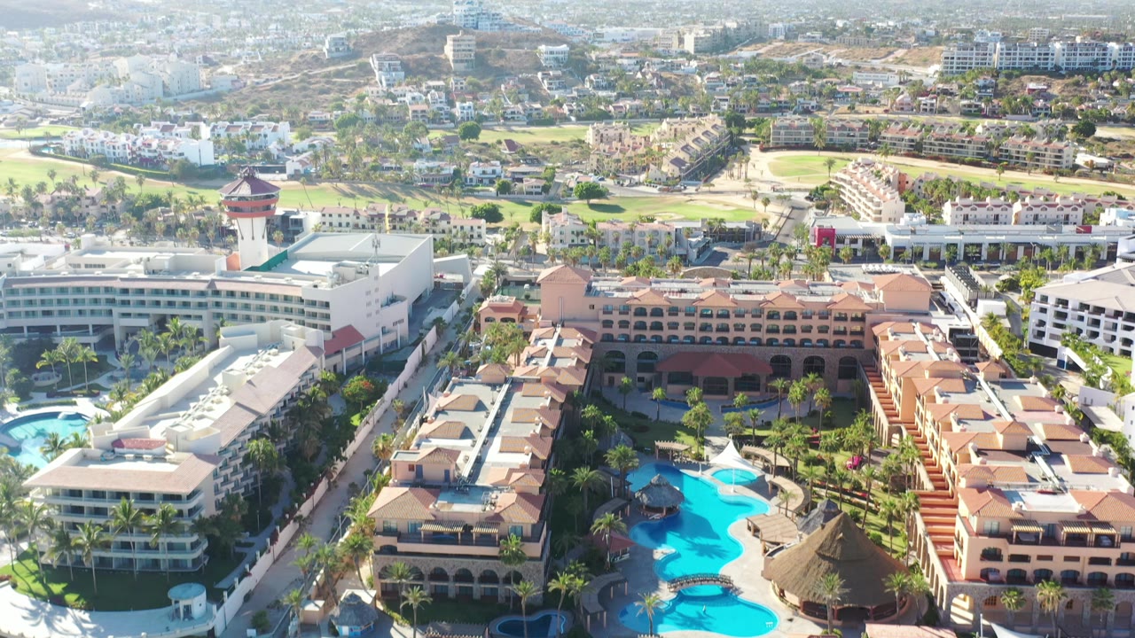 Second aerial frame in Los Cabos, Mexico, keeps the resort pools and terracotta buildings low in frame while golf fairways, hotel blocks, roads, and mountains fill the background, with no visible people.