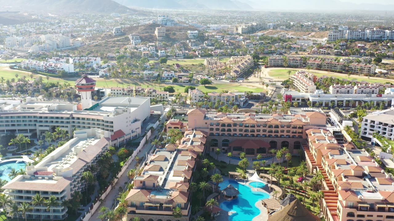Drone-height view of hotels, pools, golf course, roads, and mountain backdrop.