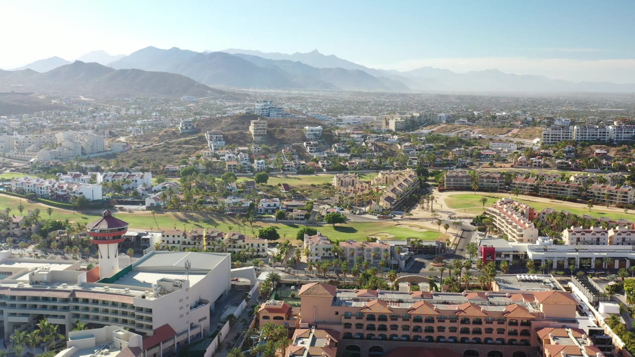 Aerial view of city buildings, golf greens, and mountains under a clear sky.