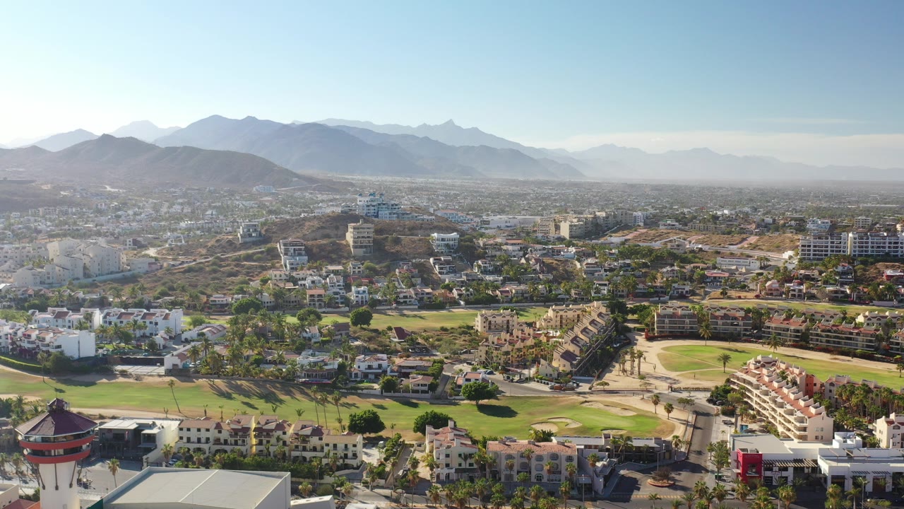 High aerial view over Los Cabos condos and white residential buildings with green grounds, roads, dry hills, distant mountains, blue sky, and no visible people.