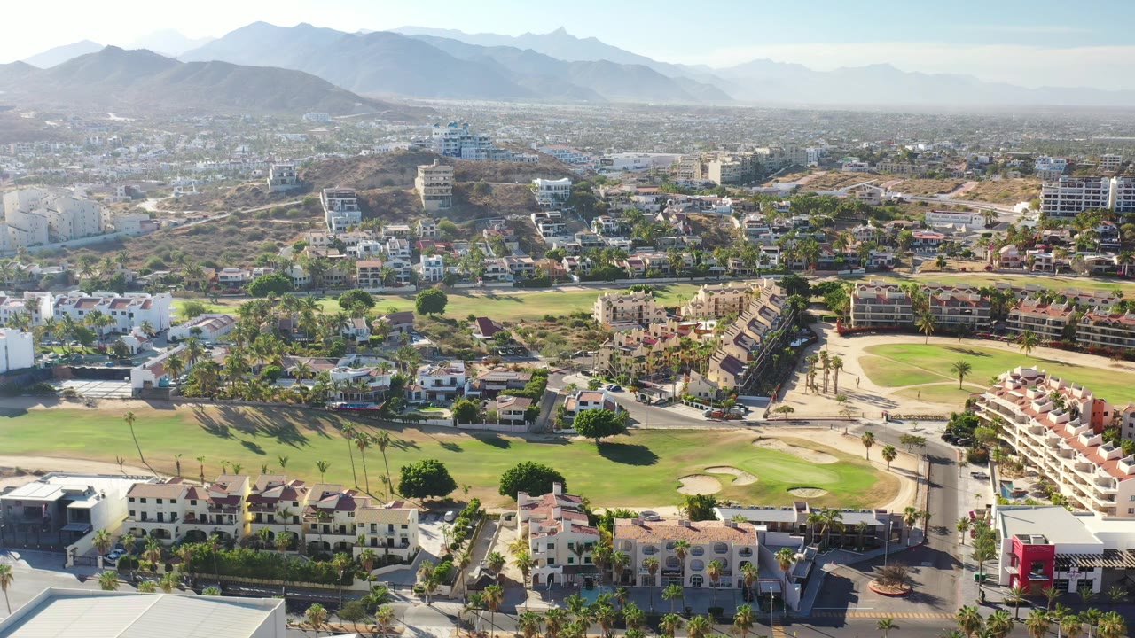 Second aerial frame in Los Cabos showing villas and apartment buildings beside green fairways, with the mountain horizon stretching across the background and no visible people.