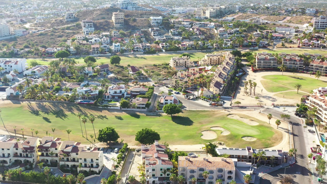 High aerial view over Los Cabos showing green golf fairways, pale sand bunkers, palm trees, roads, low-rise residences, hillside buildings, and no visible people.