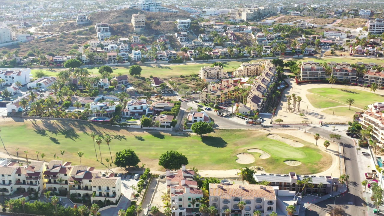 Aerial frame in Los Cabos with fairways and sand traps below, a road crossing the scene, residential buildings at the edges, hazy hillside development behind, and no visible people.
