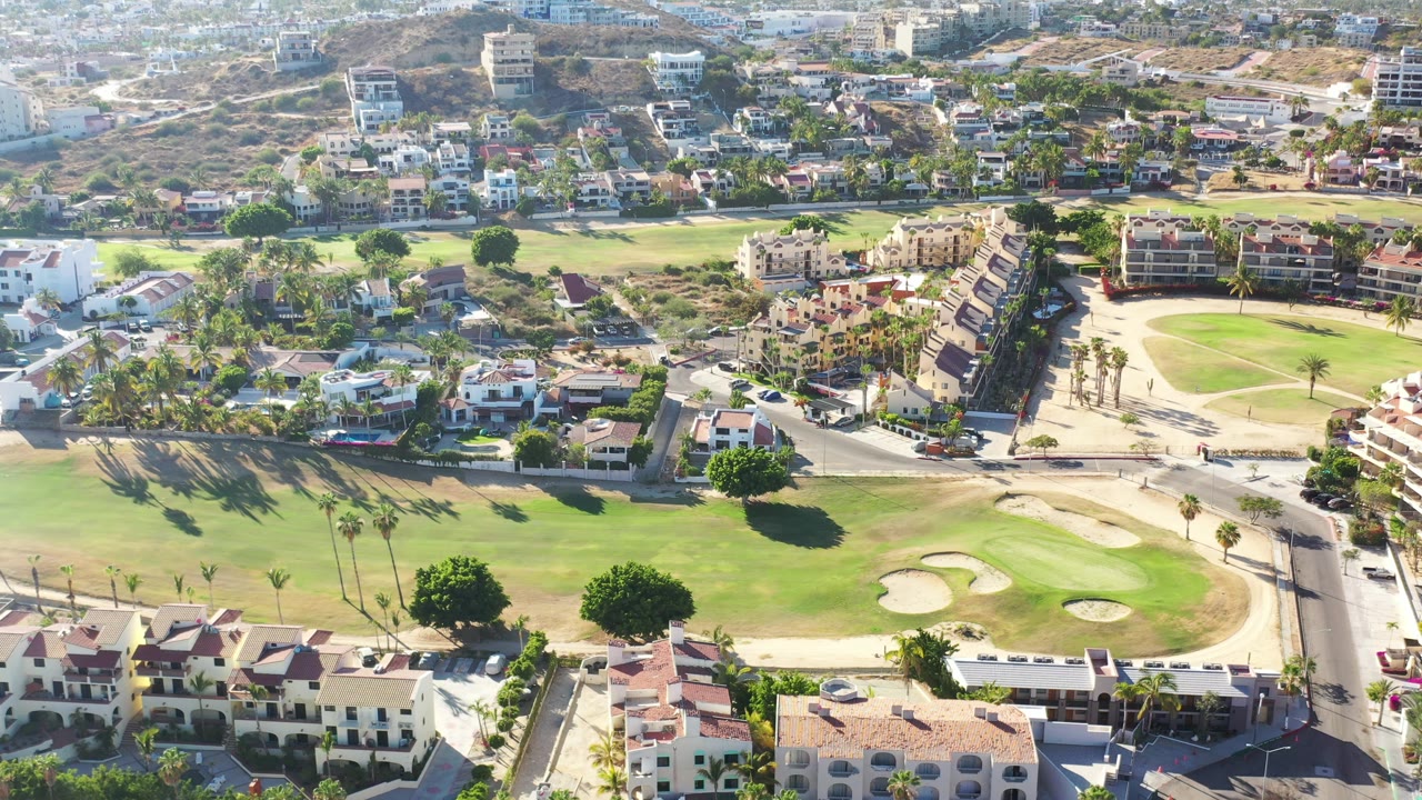 Drone view of golf greens, sand areas, palms, apartments, and hillside homes.