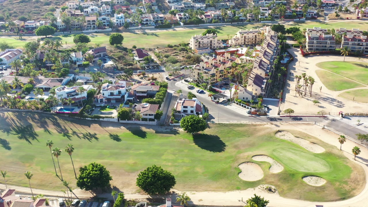 High aerial view over a Los Cabos golf community with fairway grass and sand bunkers in front, a road and homes beyond, hillside residences in the distance, and no visible people.