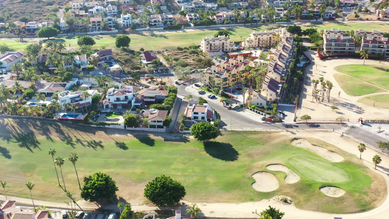 Aerial frame in Los Cabos showing a wide fairway, clustered sand traps, palm shadows, a paved road, multi-story residential buildings, and no visible people.