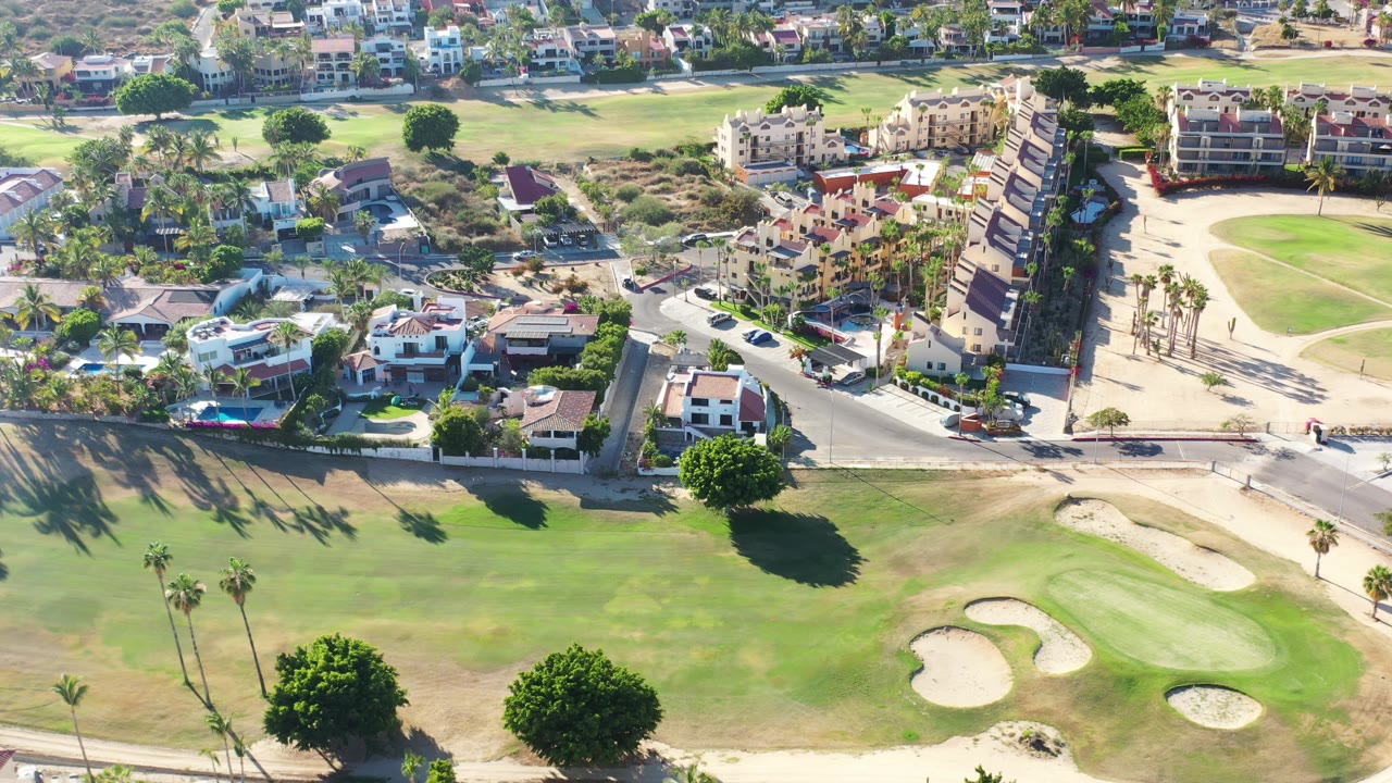 Golf green and bunkers below a road lined with homes and palm trees.