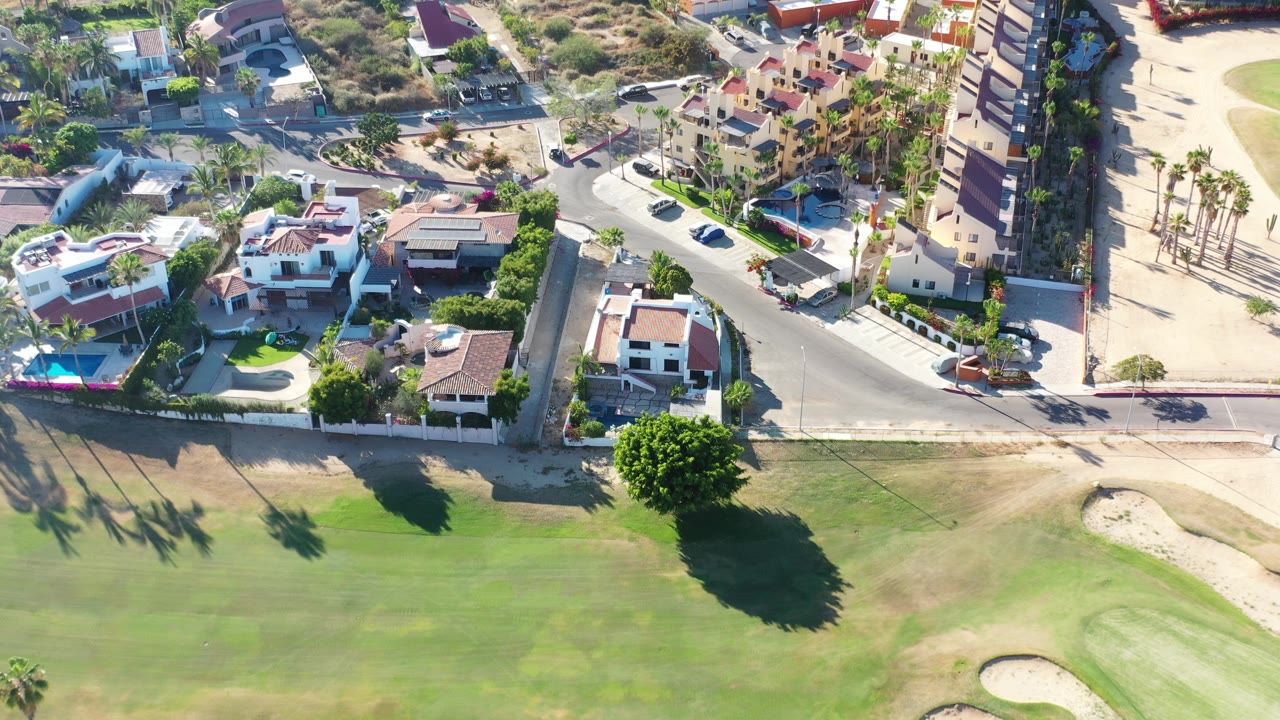 High overhead view in Los Cabos, Mexico, with resort houses, pools, a curved road, golf course grass, sand traps, palm shadows, and no visible people.