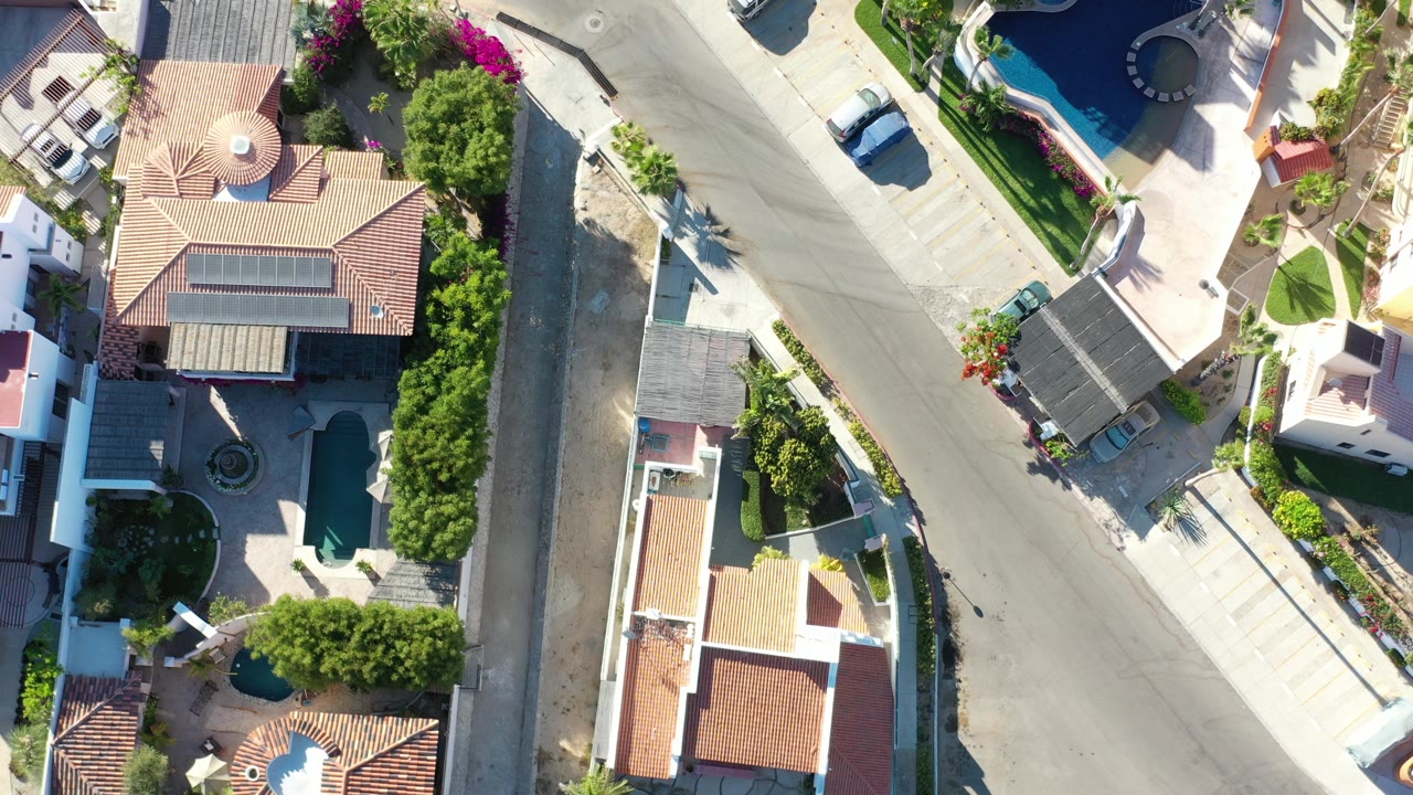 Overhead frame from Los Cabos, Mexico showing a quiet residential street corner with tile roofs, walled yards, parked cars, planted greenery, pools at the edges, and no visible people.