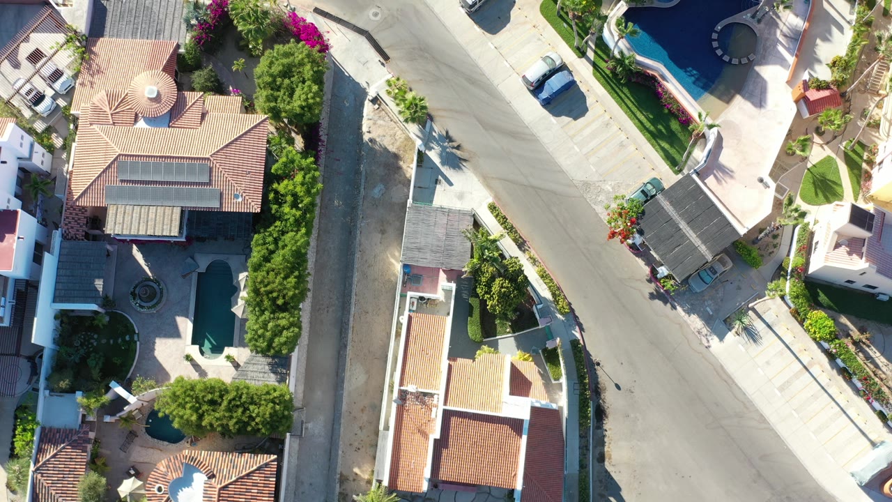 A high overhead Los Cabos view frames a curving residential road between tile-roof homes, green landscaping, a blue pool, light sidewalks, and no visible people.