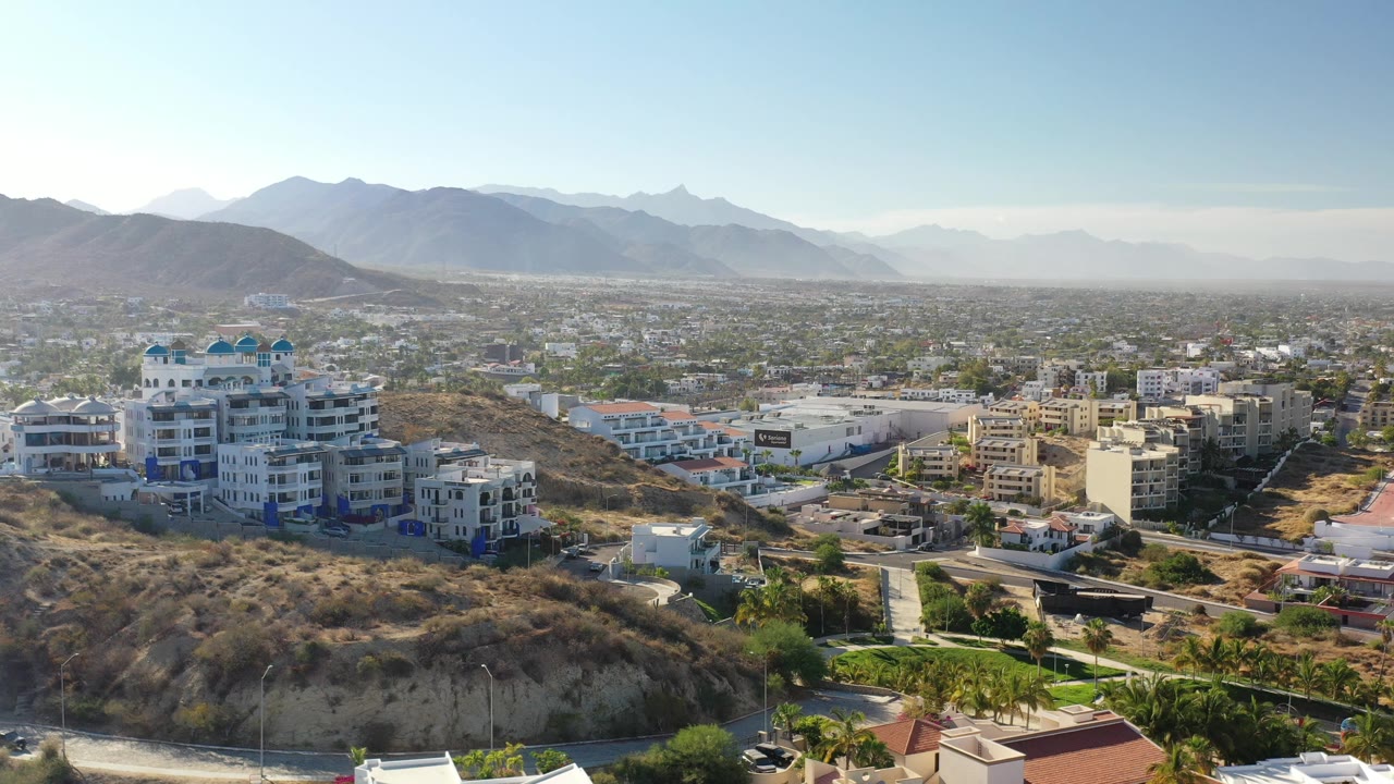 A wide aerial view over Los Cabos shows pale buildings beside a dry hillside, a spread of city blocks, hazy mountain ridges, and no visible people.