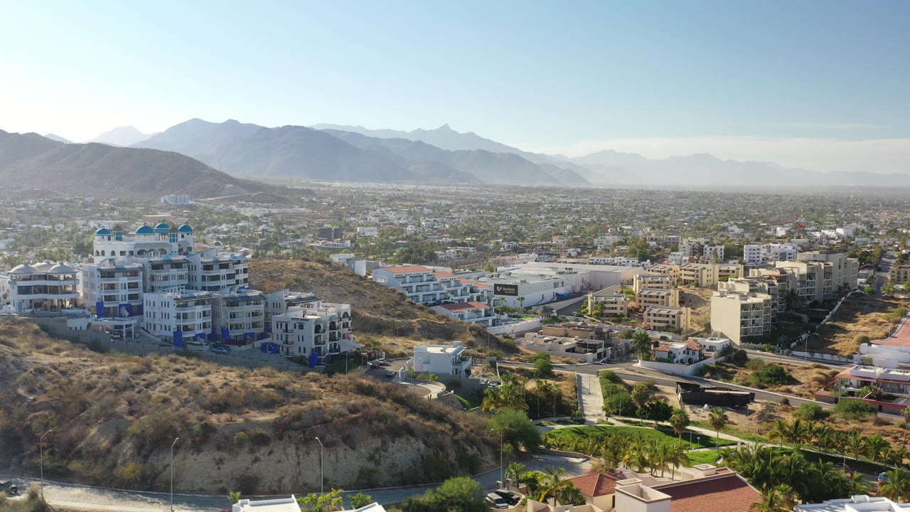 A bright Los Cabos aerial frame shows white and beige buildings at the edge of dry hills, with a large city plain and distant hazy ridges, no visible people.