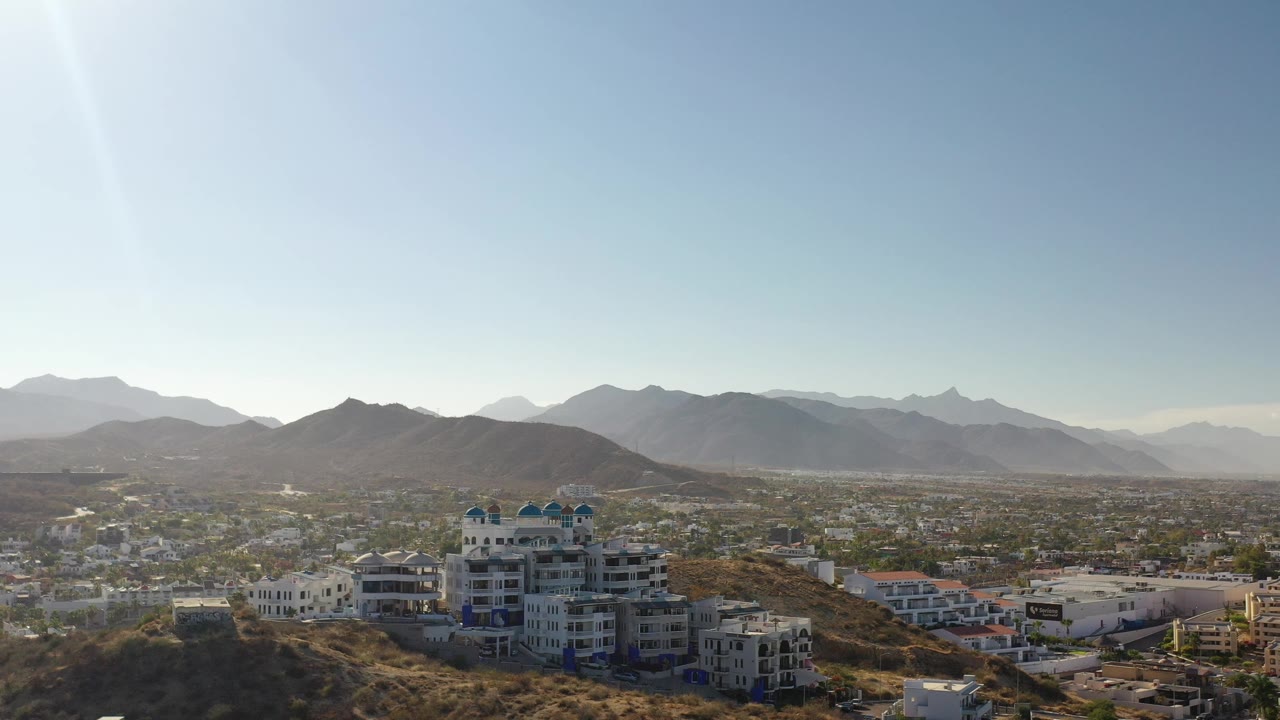 Elevated view of hillside buildings, city blocks, and distant mountains under bright sky.