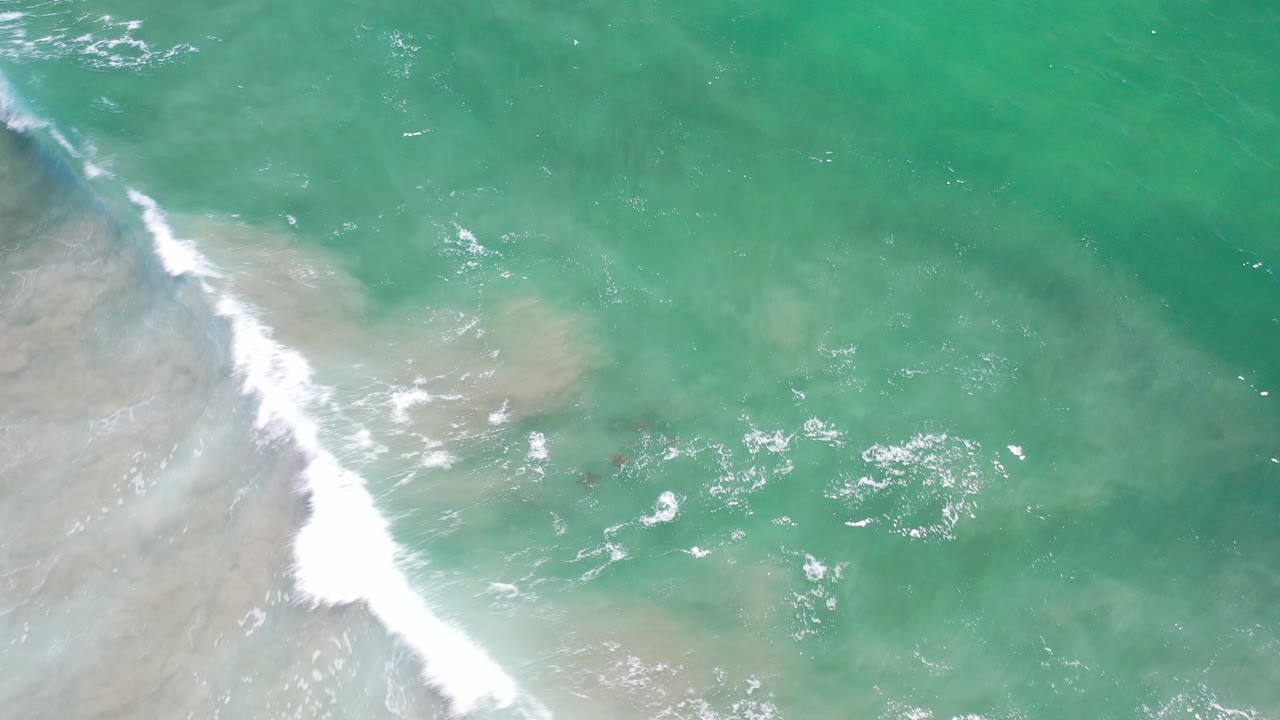 Overhead view in Los Cabos, Mexico, showing turquoise water, pale submerged sand, and white surf entering from the left with no visible people.