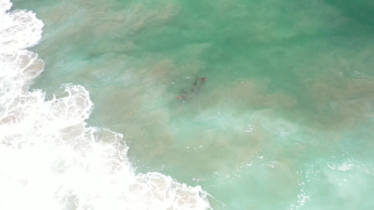 Overhead turquoise surf with white foam and tiny dark figures in the water.