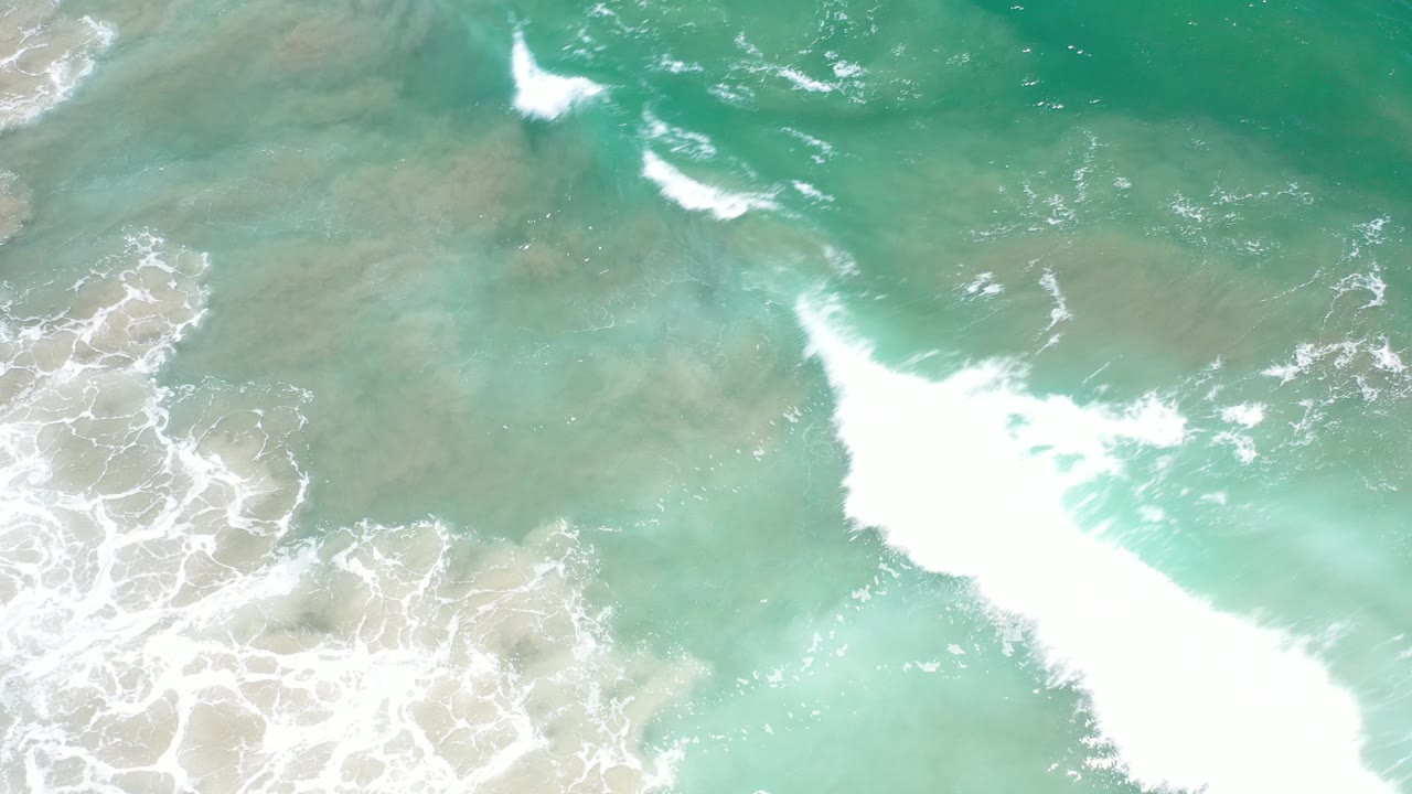 Top-down view of turquoise surf in Los Cabos, Mexico, with white foam spreading over pale shallow water and no visible people.