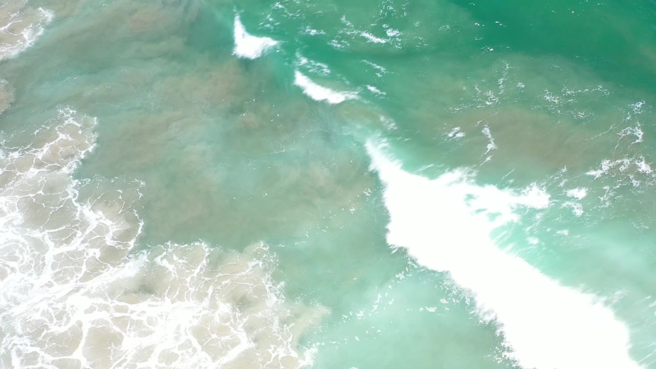 A bright white wave band crosses the overhead Los Cabos water, with pale sand visible beneath the turquoise surface and no visible people.