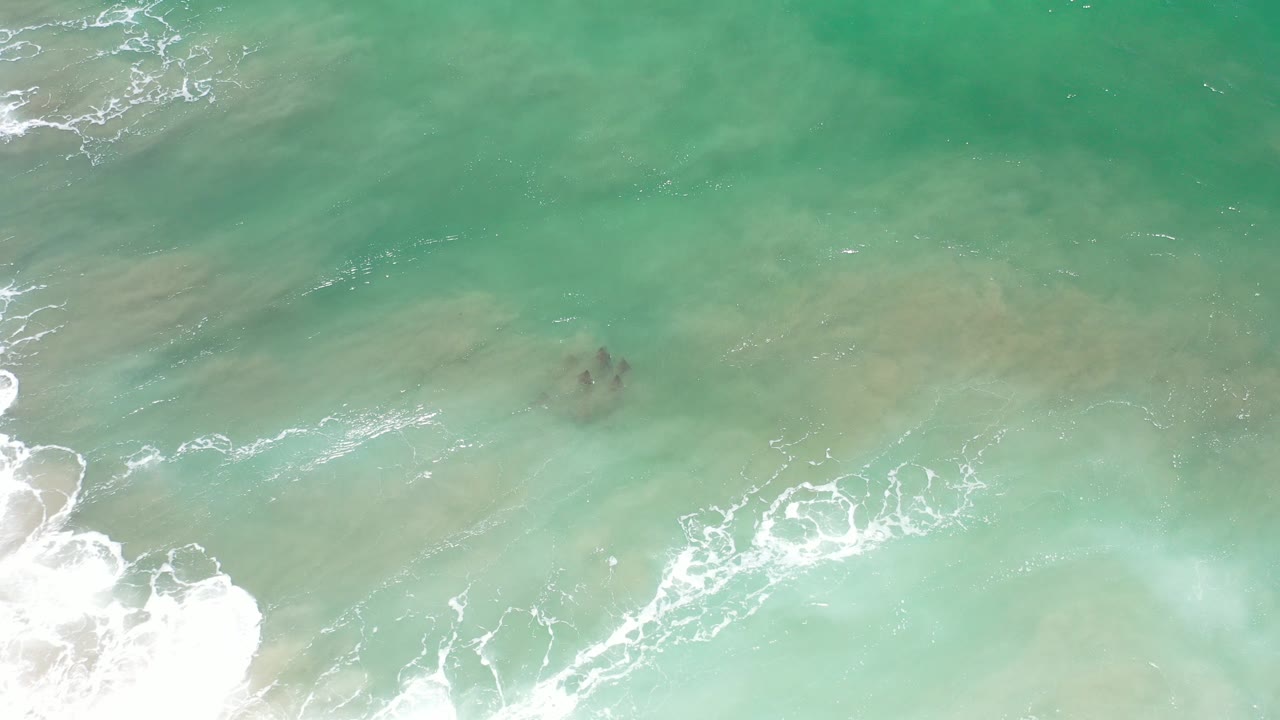 Overhead view of aqua seawater in Los Cabos, Mexico, with white foam entering from the lower left and dark submerged shapes near the center, no visible people.