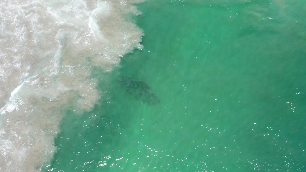 Aerial view over Los Cabos water, where pale surf hangs at the upper left and a darker underwater patch appears near center, with no visible people.