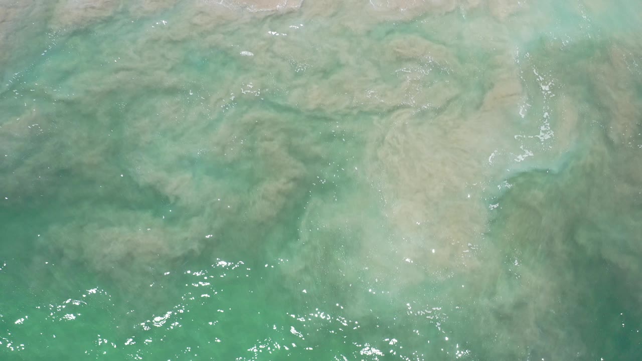 Overhead view in Los Cabos, Mexico, showing turquoise shallow ocean over tan sand before the main white wave enters, with no visible people.