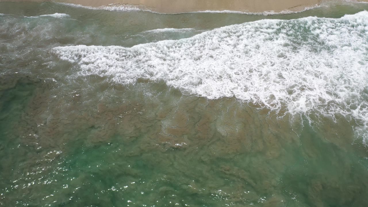A broad band of white foam crosses the shallow green water below a sandy Los Cabos shoreline, with no visible people.