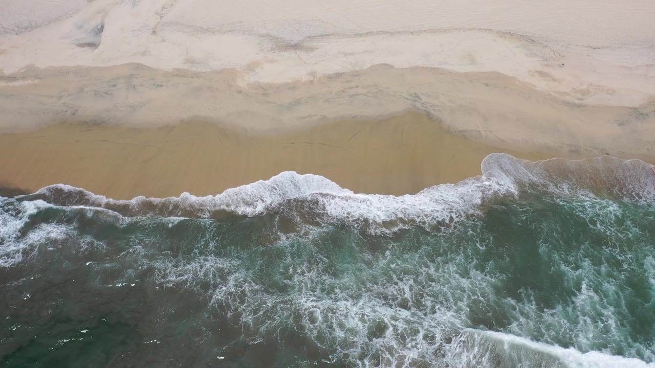 Overhead view of blue-green surf breaking onto a pale sand beach in Los Cabos, Mexico, with no visible people.
