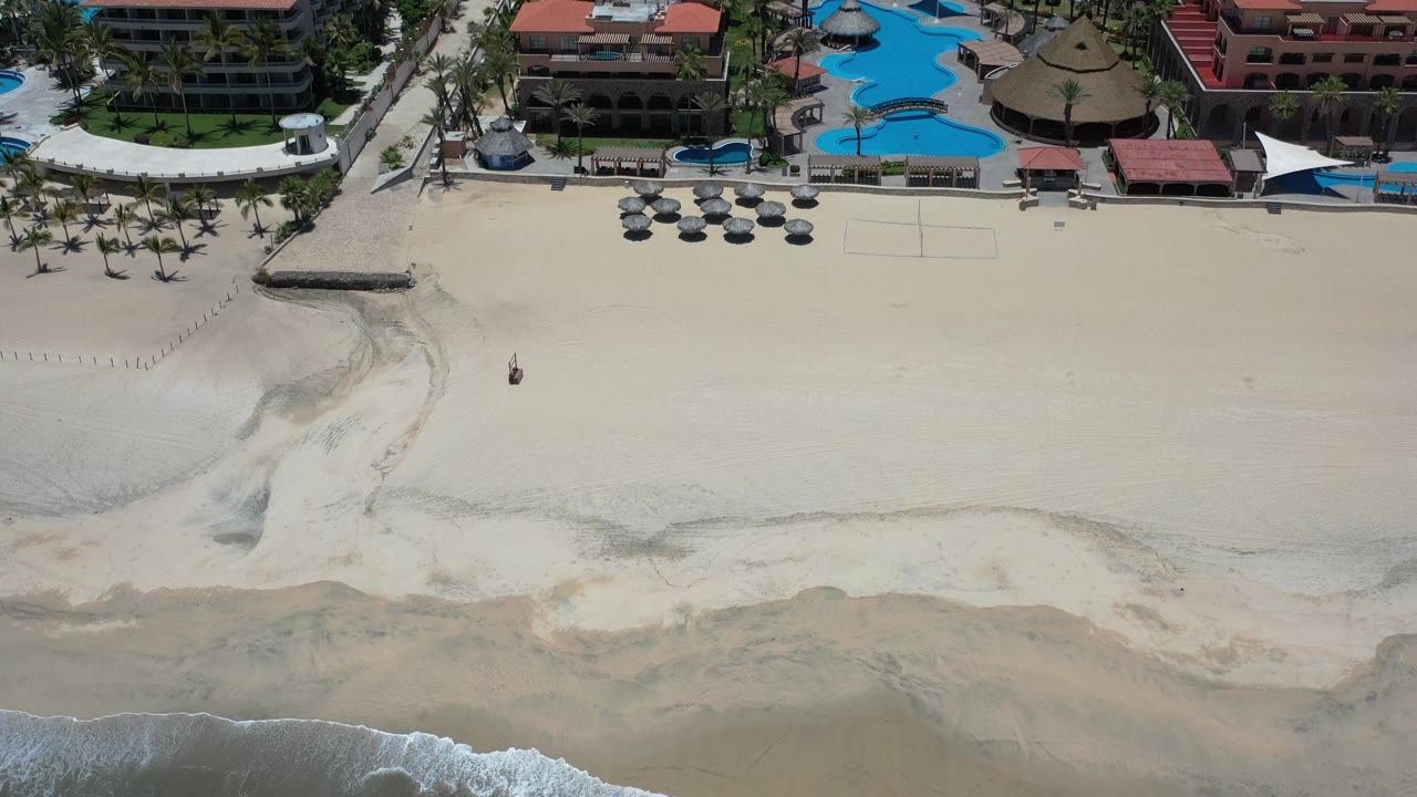 Early aerial frame over Los Cabos, Mexico, showing pale beach sand, a strip of surf, palm trees, red-roofed resort buildings, blue pools, and no visible people.