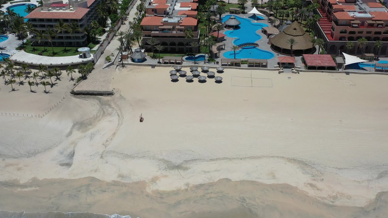 Beachfront resort with curved blue pools, umbrellas, palms, and a broad sandy beach.