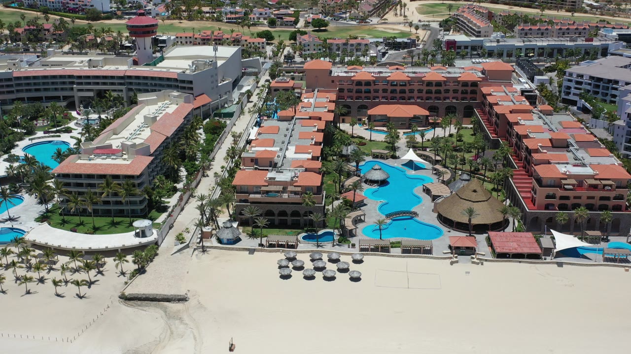 Aerial view of beachfront resort buildings, pools, palms, and sand.