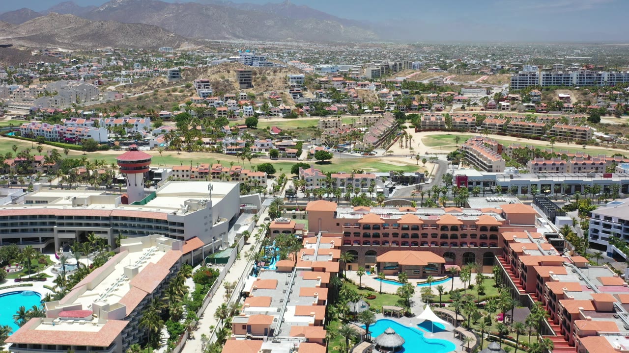 Elevated Los Cabos frame showing hotel rooftops and pools at the bottom, a road and golf-course fairways through the center, spread-out buildings, hazy mountains, and no visible people.