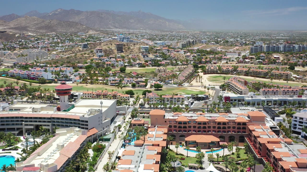 Aerial Los Cabos view with a white hotel block, orange-roofed resort wings, pools, palm-lined grounds, urban buildings across the valley, arid hills, mountains, and no visible people.