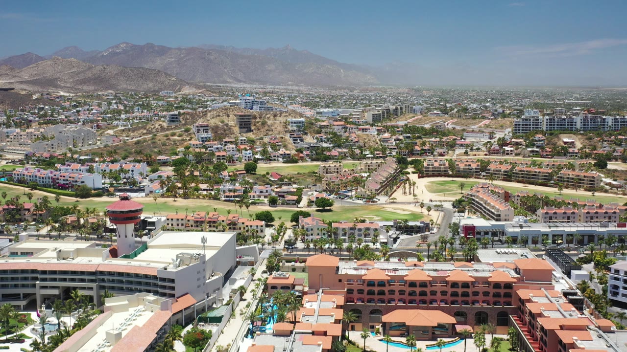 Wide overhead view of Los Cabos buildings, golf greens, hills, and resort rooftops.