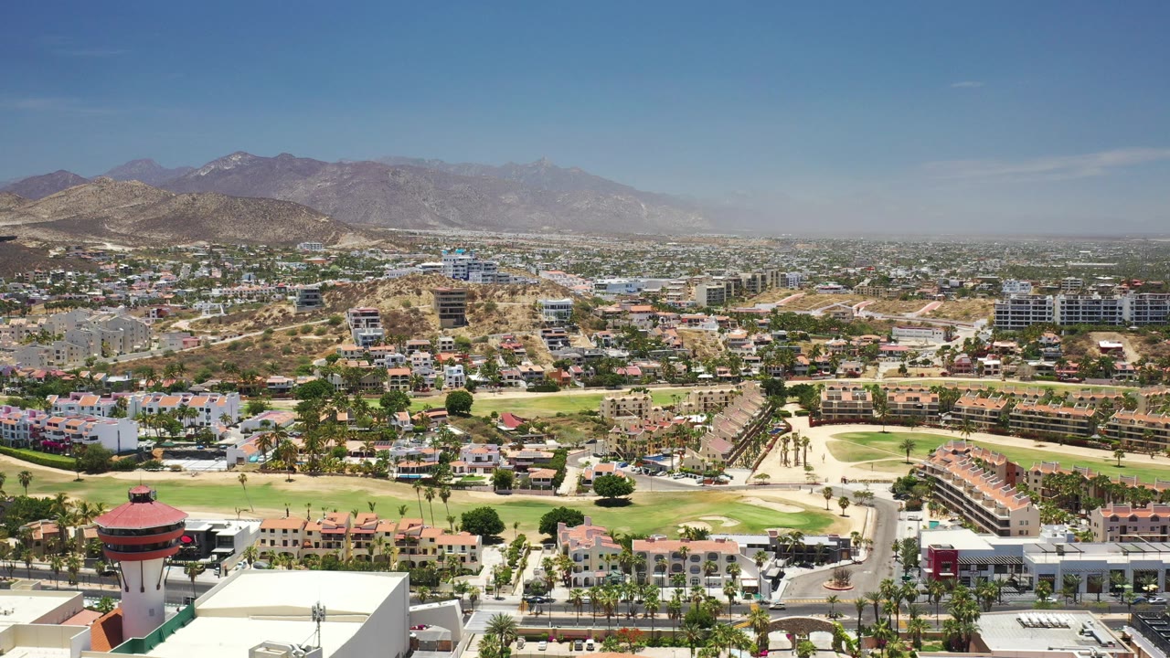 Wide aerial view over Los Cabos, Mexico, with roads, red-roofed buildings, green fairways, tan hills, distant mountains, and no visible people.