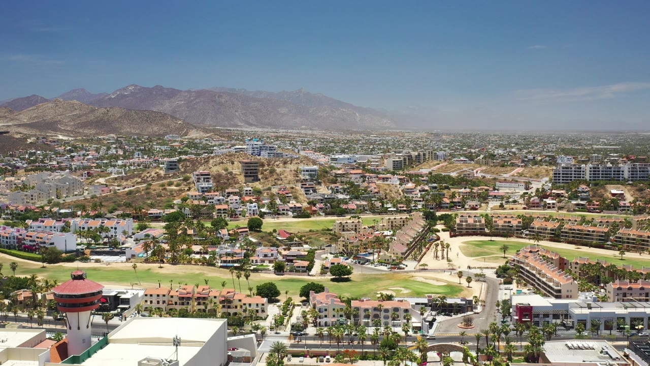 Aerial frame over Los Cabos showing a red-topped tower near the foreground, clustered buildings, green fairways, dry hills, distant mountains, and no visible people.