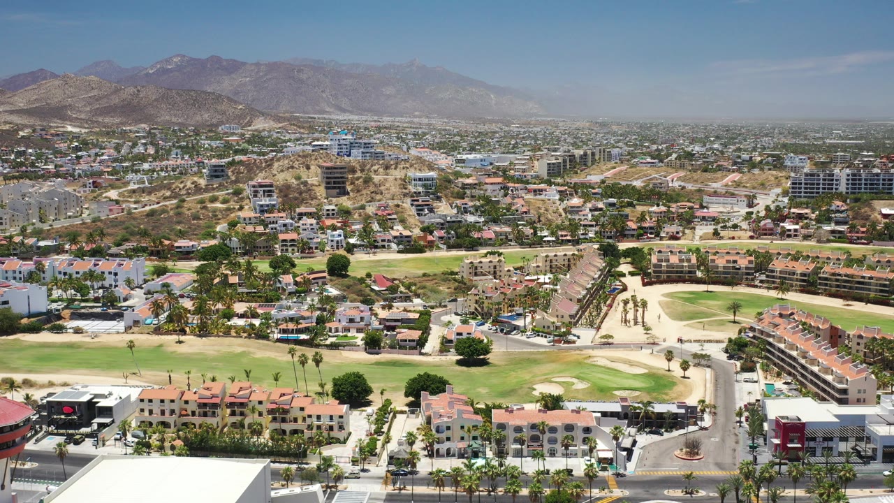 Opening aerial frame over Los Cabos, Mexico, showing red-roofed homes, pale golf-course fairways, roads, low-rise buildings, desert mountains, and no visible people.