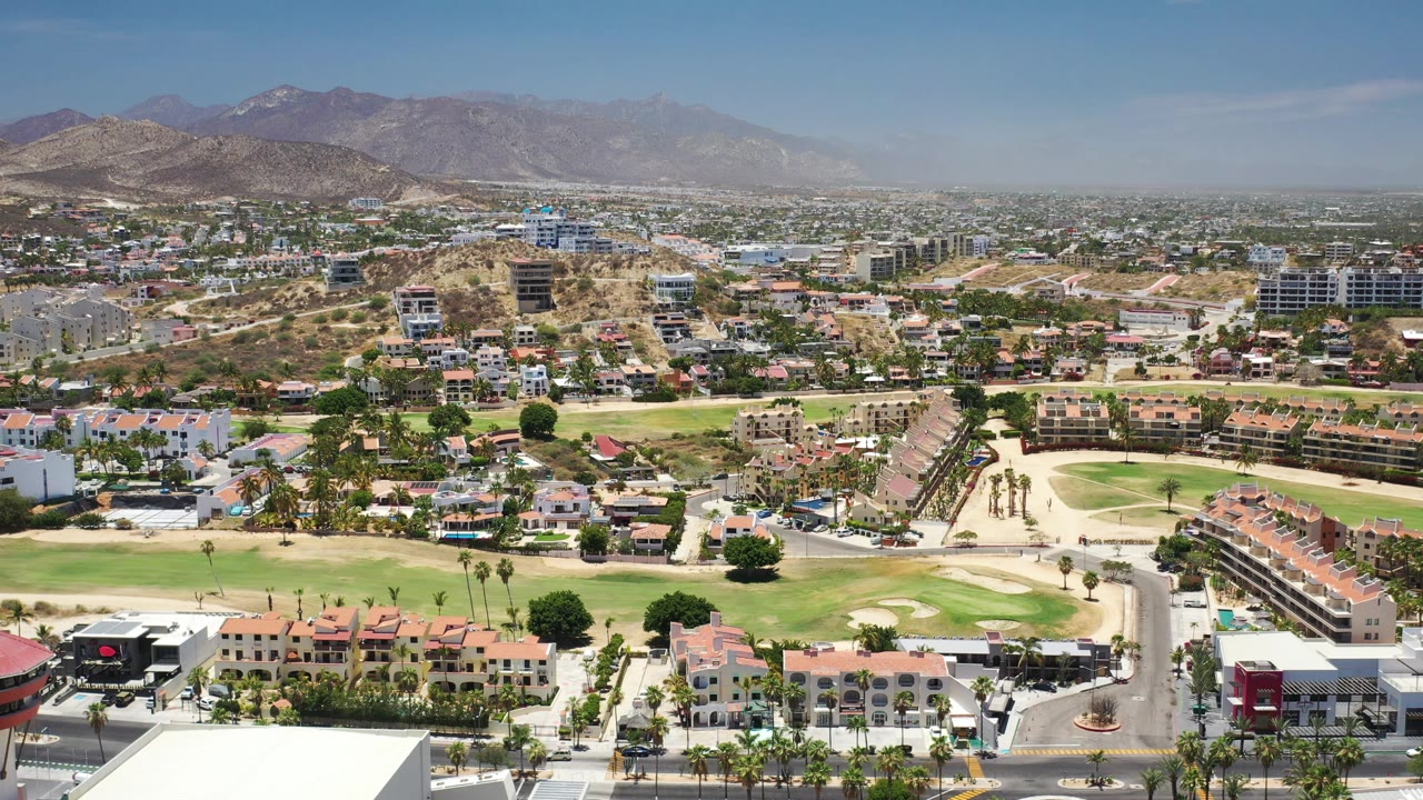 Second aerial frame in Los Cabos, Mexico, with foreground rooftops, a central golf-course green, apartment buildings, hillside homes, hazy mountains, and no visible people.