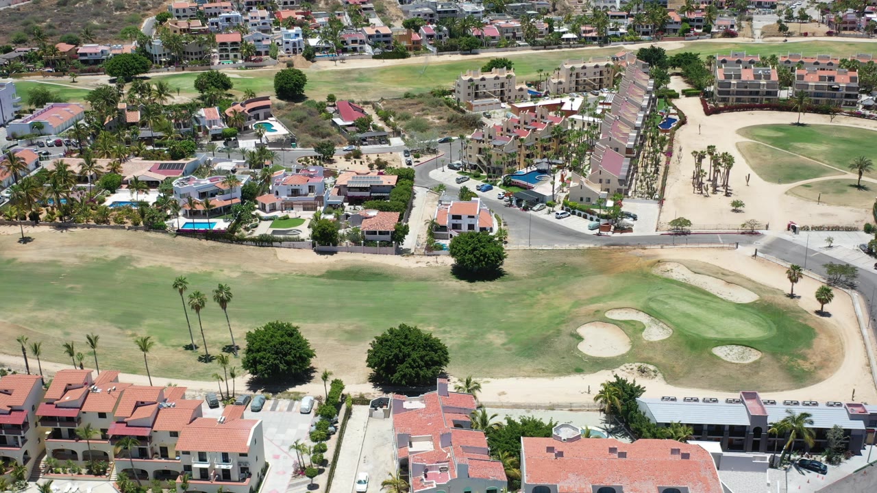 Aerial frame over Los Cabos, Mexico showing red-tile buildings, parked cars, palm trees, a road, and green golf-course turf with sand bunkers, with no discernible people.