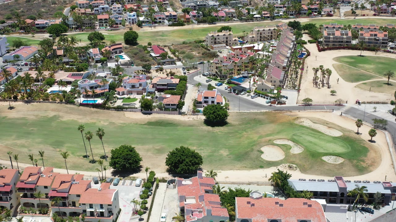 Los Cabos aerial view with residential buildings at the lower edge, a paved road through the center, palms, dry lots, and rounded golf-course sand bunkers in bright sun, with no discernible people.
