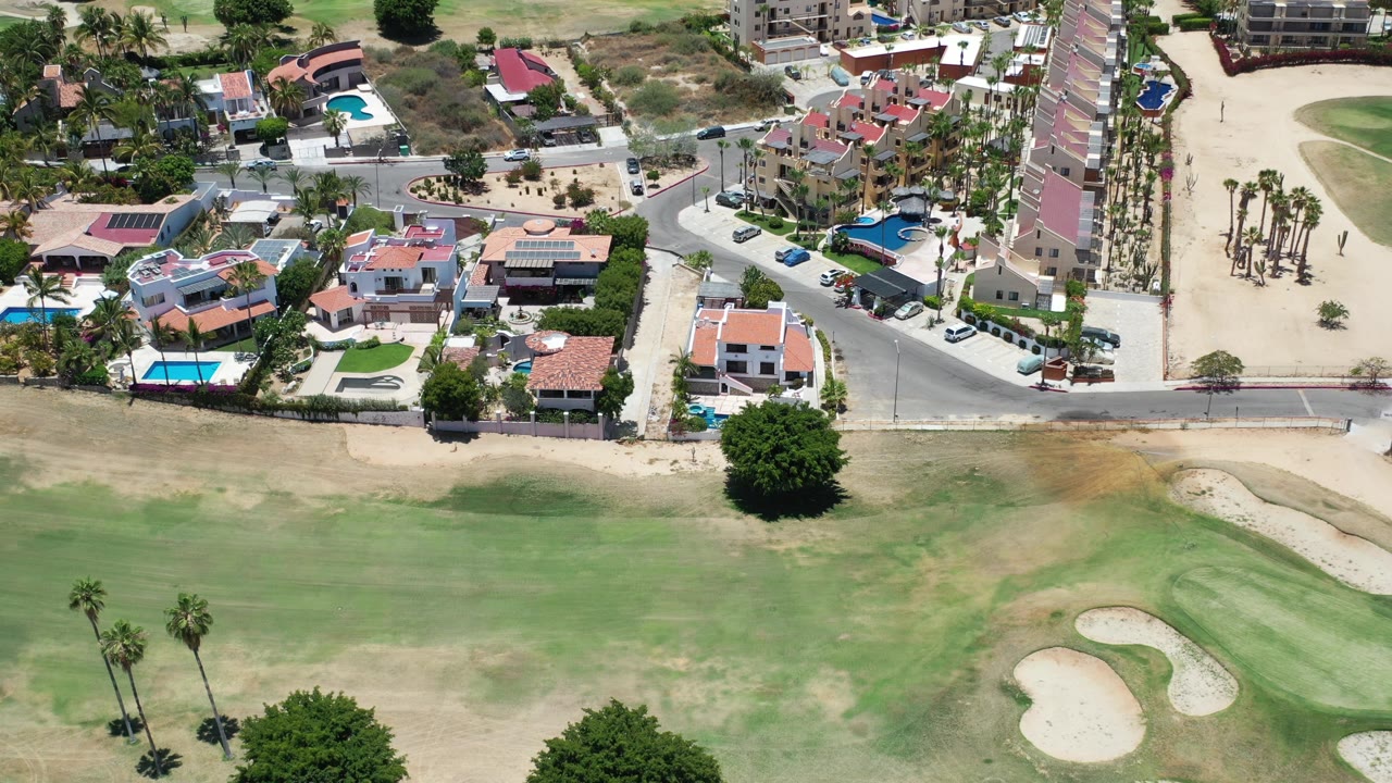 High aerial view in Los Cabos, Mexico, showing residential homes with pools beside a curved street and golf-course grass in the foreground, with tiny distant people near the sandy area.