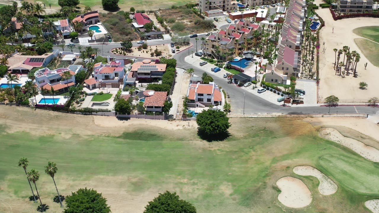 Aerial frame over Los Cabos homes with red tile roofs, blue pools, palm-lined paths, a paved road, and green fairway grass filling the lower half of the view.